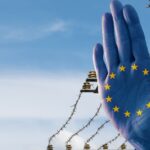 A blue hand with the European Union stars is raised in front of barbed wire, symbolizing protection or barriers under a clear sky.