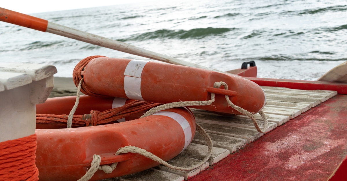 Two orange life rings with white stripes rest on a red boat near the shore, with ocean waves in the background.