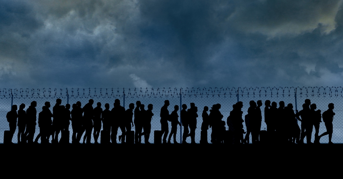 Silhouettes of people standing in line near a barbed wire fence under a dark, cloudy sky, suggesting a border or refugee scene.