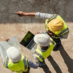 Three construction workers in safety gear reviewing plans and a laptop on a building site.