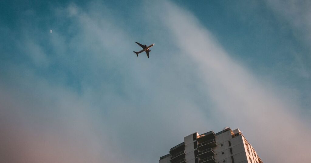 An airplane flies above a tall city building against a blue sky with soft clouds.