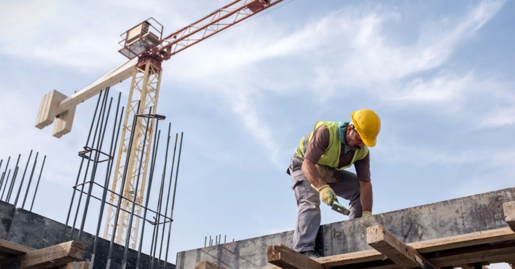 A construction worker in a hard hat and vest works on a concrete structure with a crane in the background.