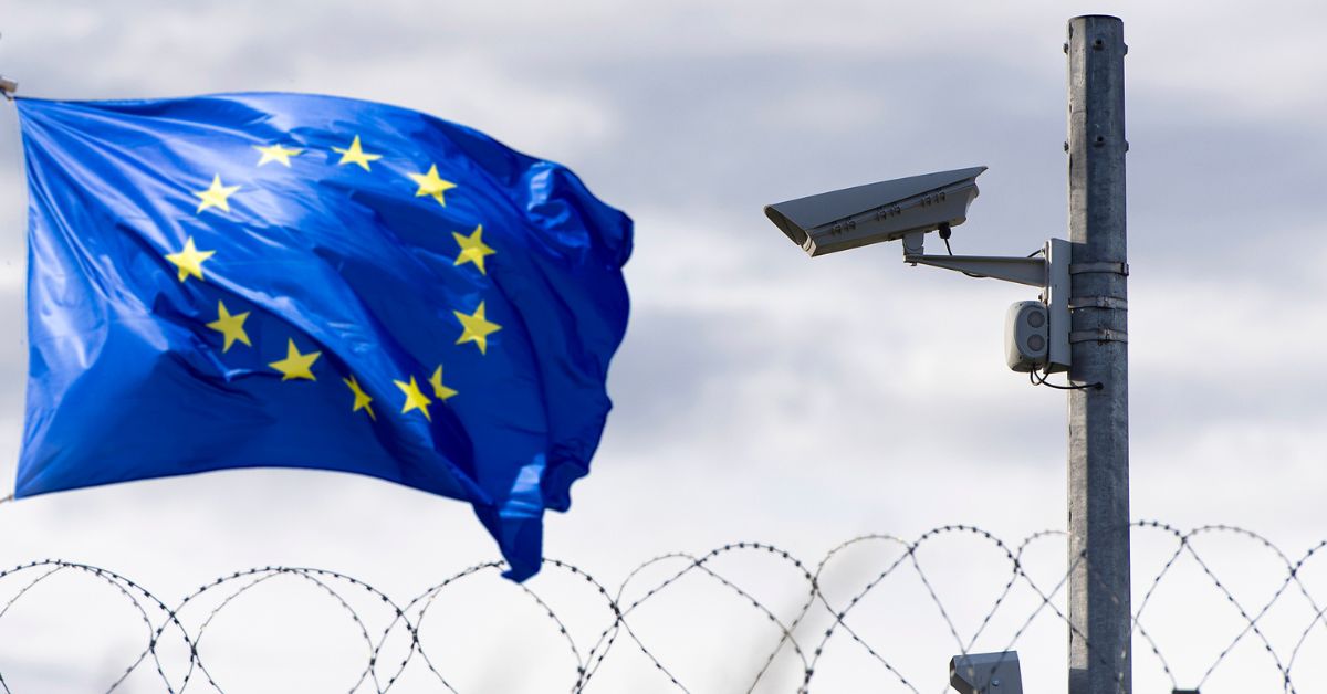 EU flag waves near a security camera above barbed wire at a border.
