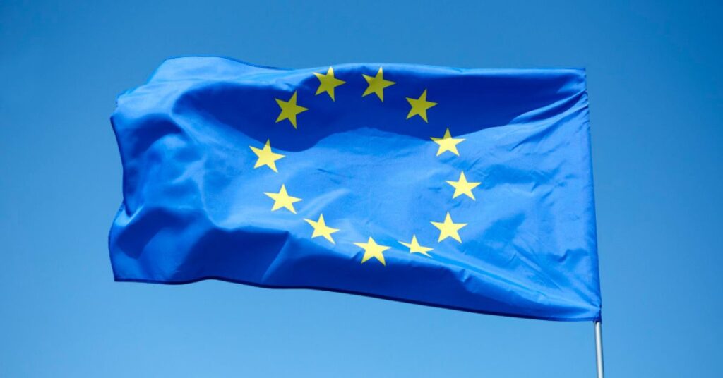 The European Union flag with yellow stars in a circle on a blue background, waving against a clear sky.