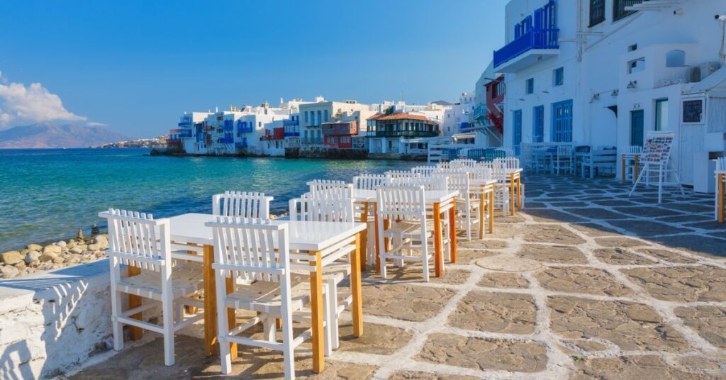Seaside cafe with white tables and chairs overlooking colorful buildings by the blue sea in the Greek islands.