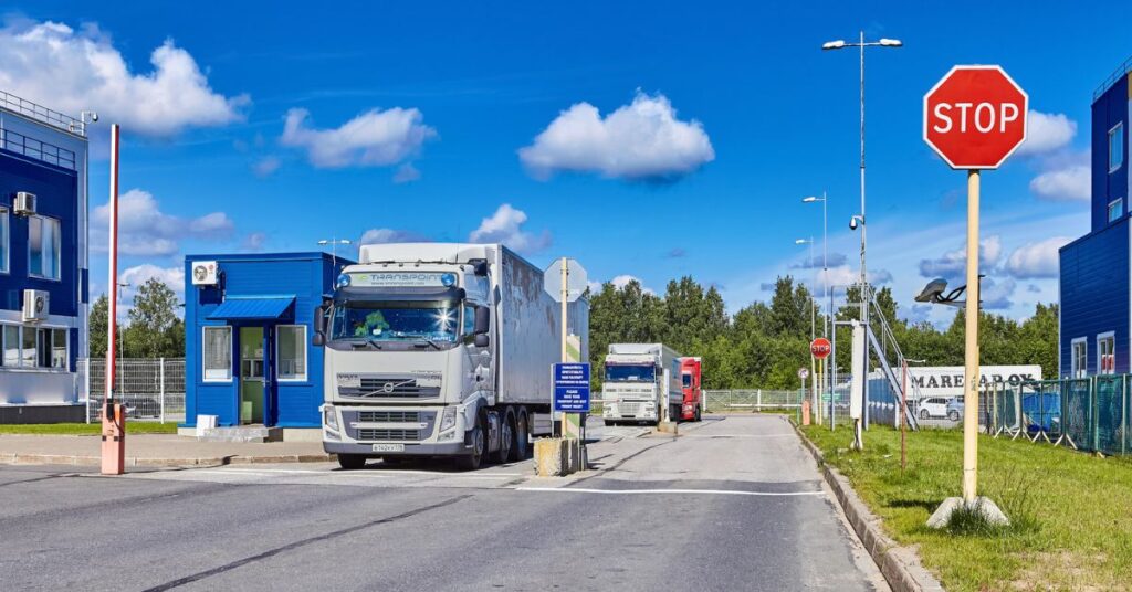 Several trucks lined up at a border checkpoint with stop signs, guard booths, and a clear blue sky overhead.