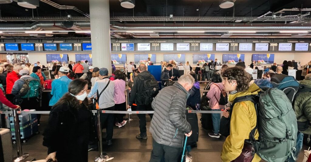 Travelers stand in long lines at airport check-in counters with luggage and backpacks.