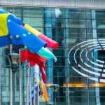 Flags of EU member states and Ukraine fly outside the European Parliament building with the EU emblem on the glass facade.