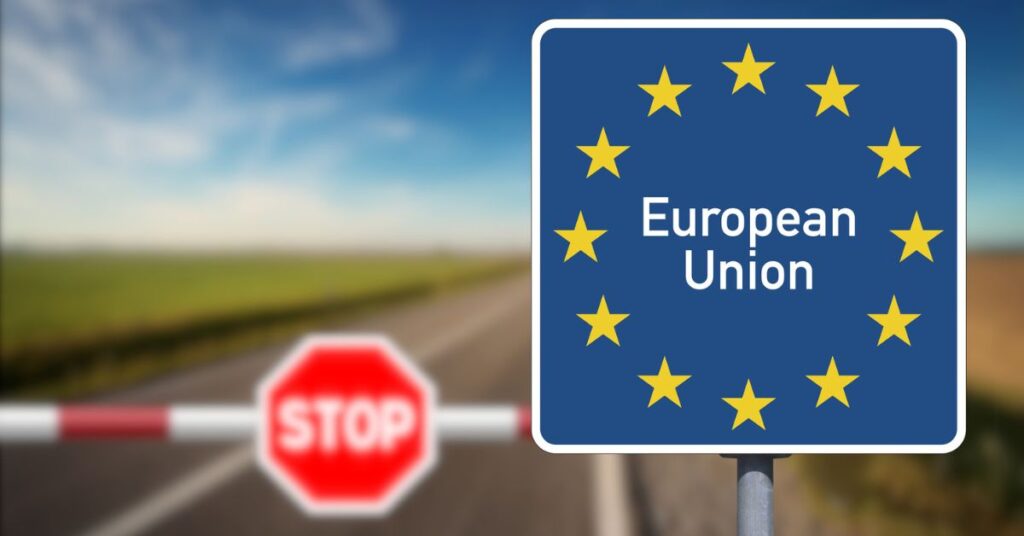 A road sign with the European Union flag and text stands beside a red stop barrier on a rural road.