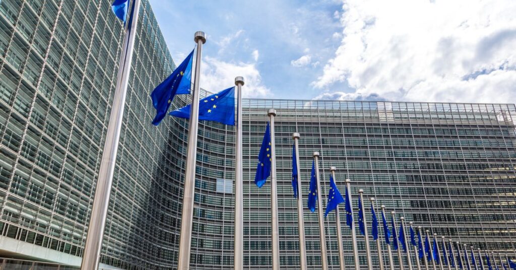 Row of European Union flags waving outside a large glass-fronted office building under a partly cloudy sky.