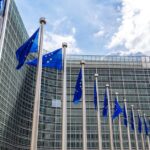 Row of European Union flags waving outside a large glass-fronted office building under a partly cloudy sky.