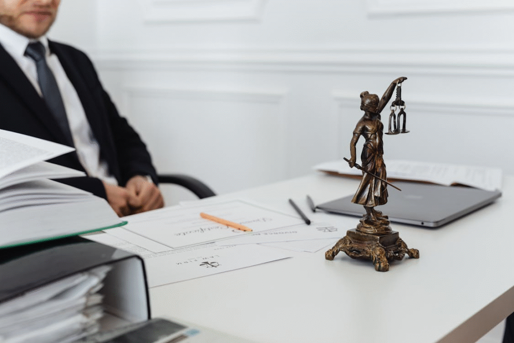 A lawyer sits at a desk with documents, a laptop, and a Lady Justice statue in the foreground.