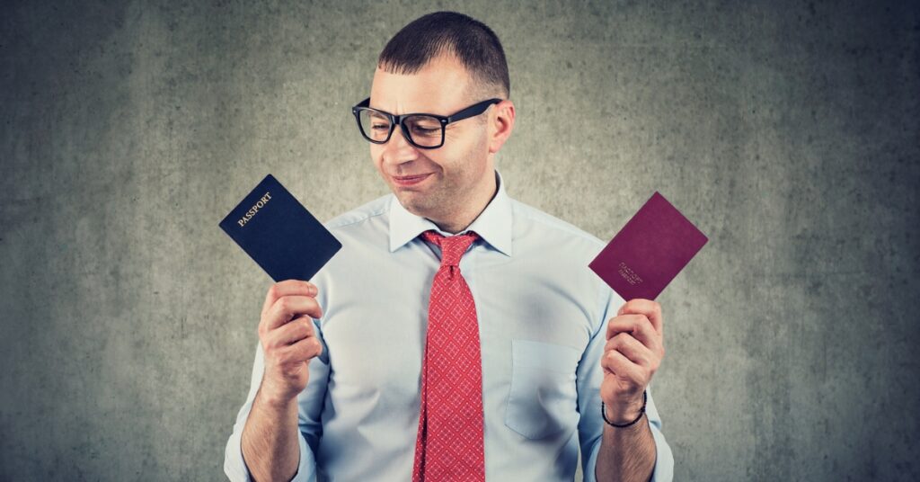 Man in glasses and red tie holding two passports, looking down at them with a thoughtful expression.