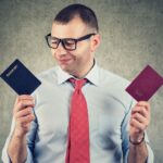 Man in glasses and red tie holding two passports, looking down at them with a thoughtful expression.