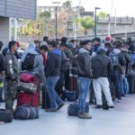 A large group of people with backpacks and suitcases stand in a long outdoor line.