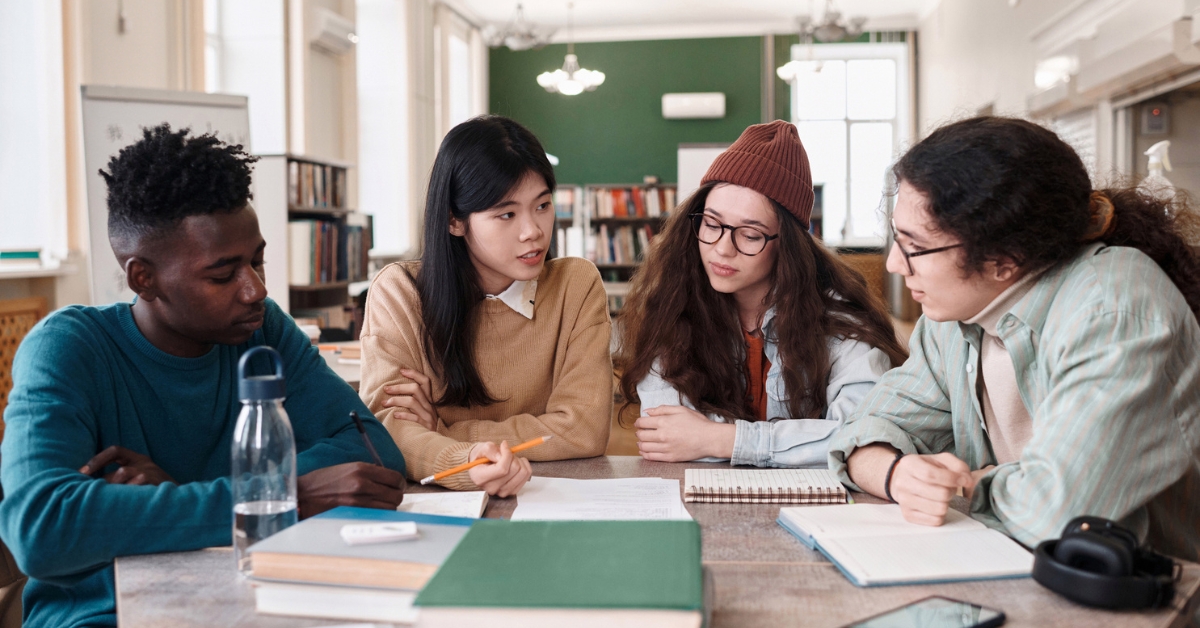 Four students sit at a library table with books and notebooks, discussing and working together on their studies.