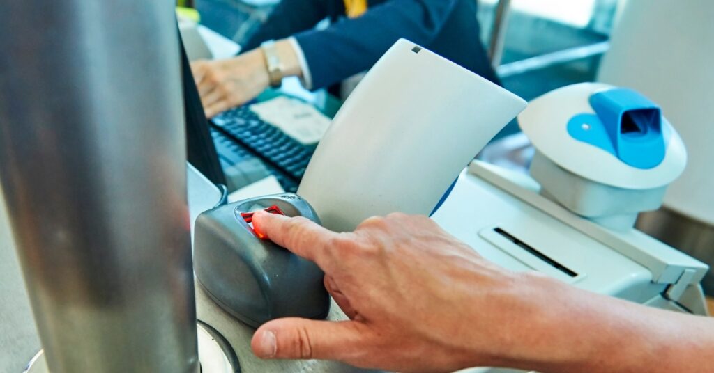 Traveler places a finger on a biometric scanner at an airport security checkpoint while an officer works at a computer.