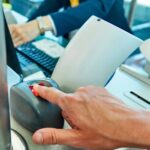 Traveler places a finger on a biometric scanner at an airport security checkpoint while an officer works at a computer.