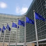 Rows of European Union flags wave in front of a large glass office building under a blue sky.