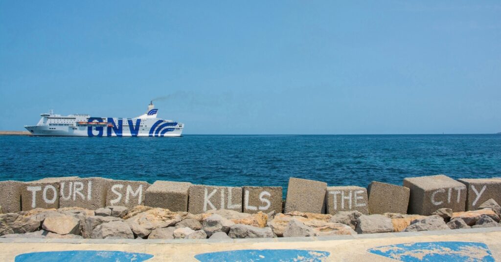 Ferry sailing past a seawall with graffiti reading “Tourism kills the city” under a clear blue sky.