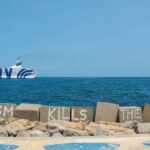 Ferry sailing past a seawall with graffiti reading “Tourism kills the city” under a clear blue sky.