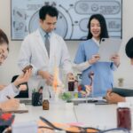 Medical students in lab coats and scrubs studying anatomy models and medications in a classroom setting.