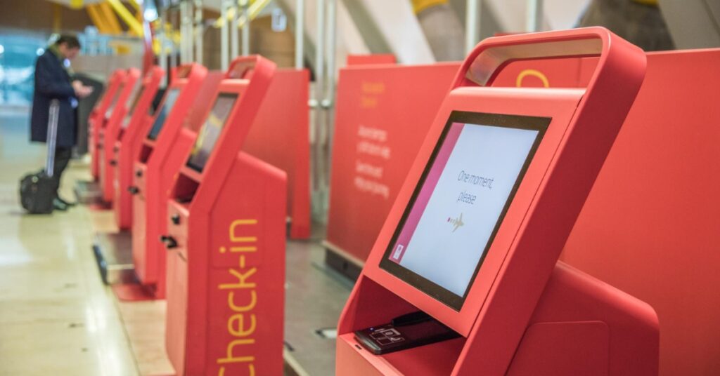 A row of red airport self check-in kiosks displays a loading message while a traveler waits nearby with luggage.