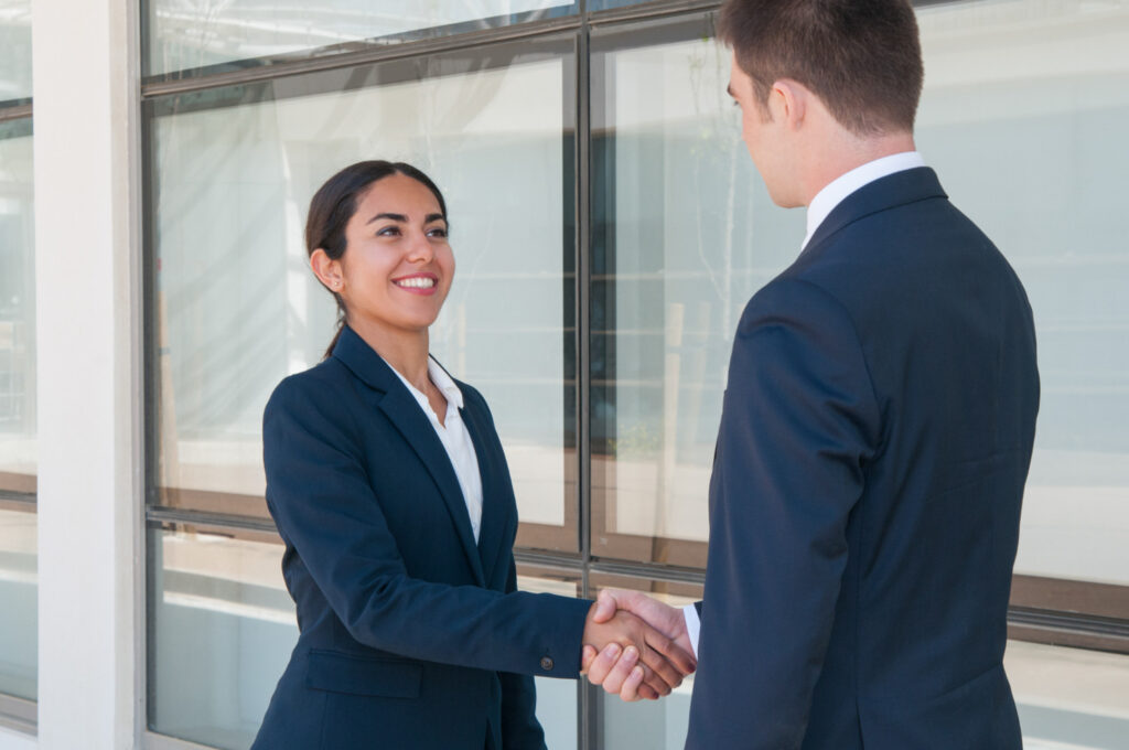 Two business professionals in suits shake hands outside a modern building, smiling warmly at each other.