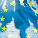 Silhouetted people cast long shadows across pavement with the translucent blue and yellow stars of the EU flag overlayed on the scene.