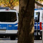 Two German police vans with blue markings are parked on a shaded street lined with trees.