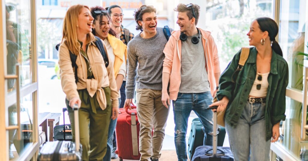 A group of young adults laughing as they walk into a building with rolling suitcases.