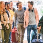 A group of young adults laughing as they walk into a building with rolling suitcases.