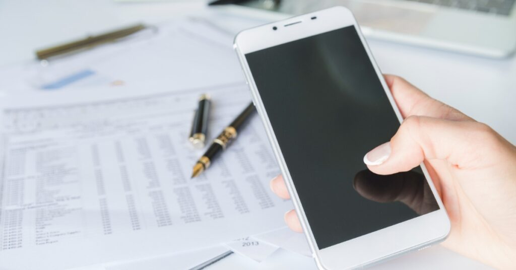 A hand holds a smartphone above financial documents and a pen on a bright desk.
