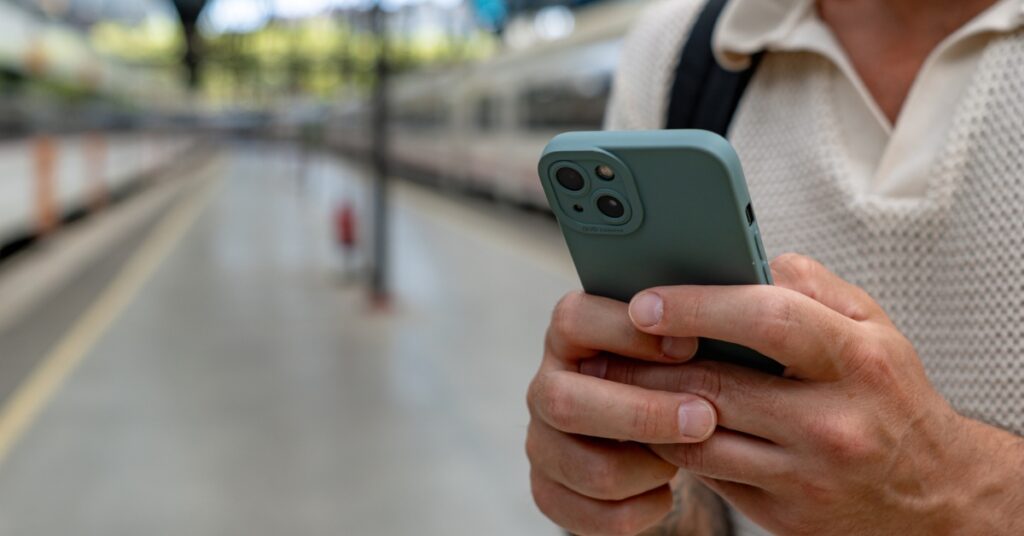 A man holding a smartphone while standing on a train station platform with blurred trains in the background.