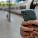 A man holding a smartphone while standing on a train station platform with blurred trains in the background.