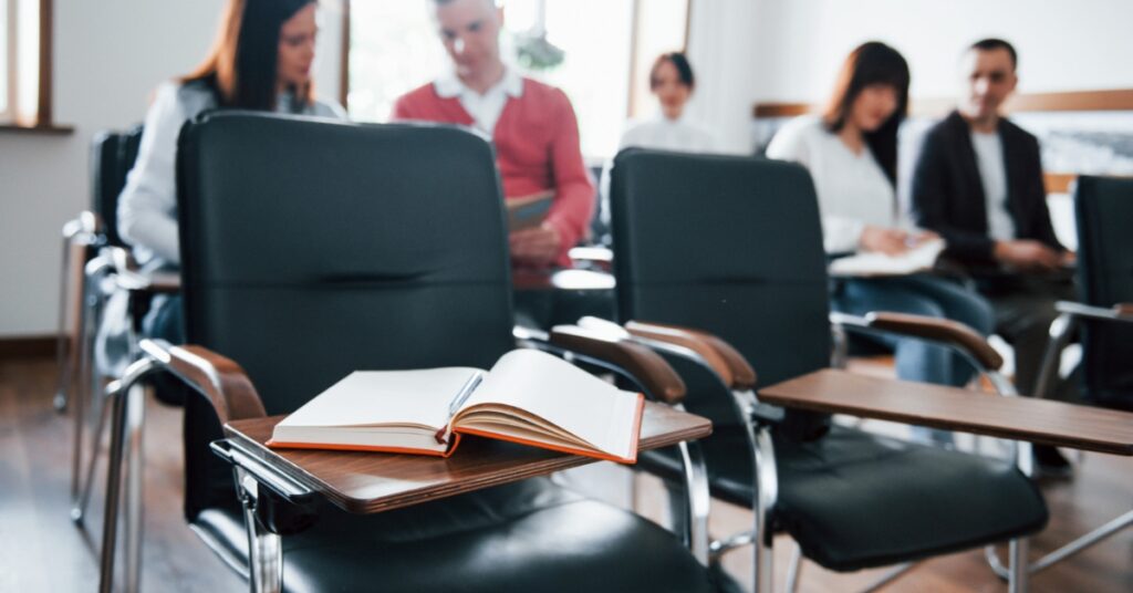An open notebook rests on a classroom chair’s desk arm as students sit and read in the background.