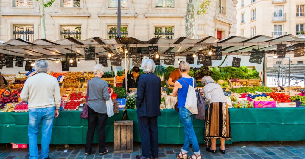 People browse fresh fruits and vegetables at an outdoor farmers market in a city square.