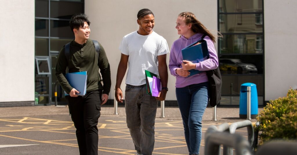 Three students walk together outside a campus building, smiling and carrying notebooks and folders.