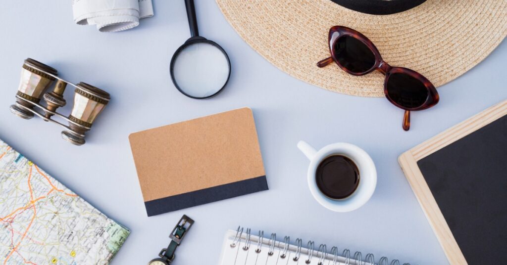 A flat lay of travel items including a hat, sunglasses, map, notebook, coffee, magnifying glass, and binoculars.
