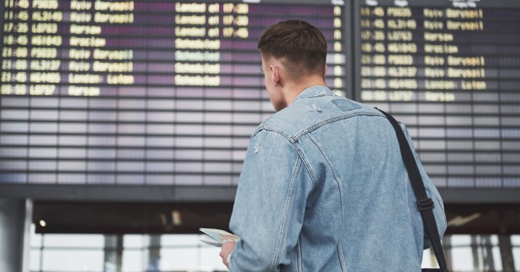 A man in a denim jacket looks up at a large airport departures board while holding travel documents.