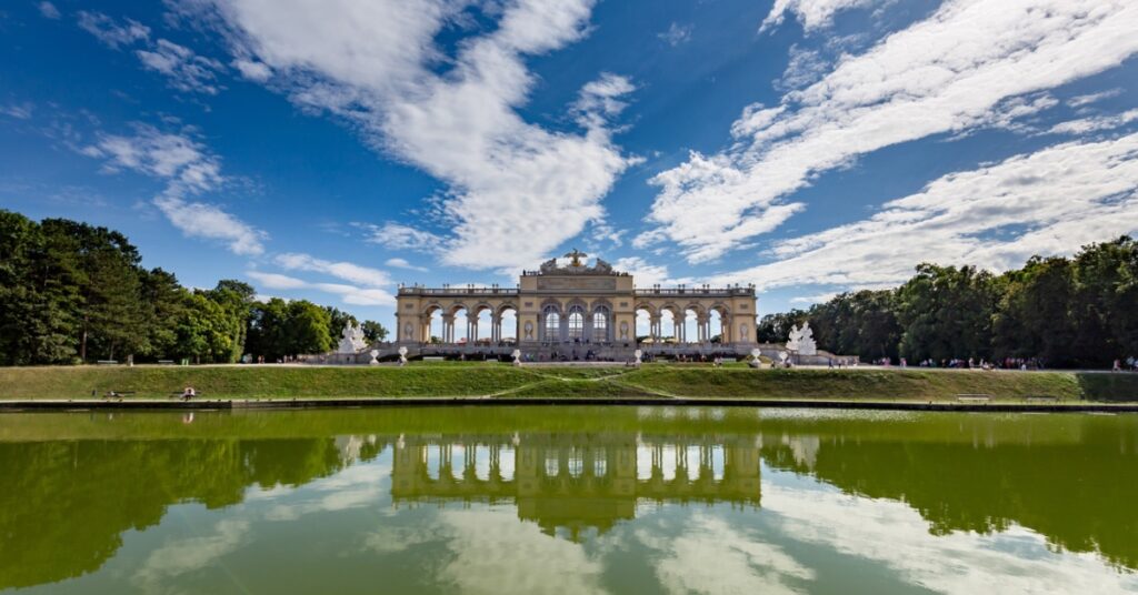 The Gloriette at Schönbrunn Palace stands reflected in a calm lake under a bright sky with scattered clouds.