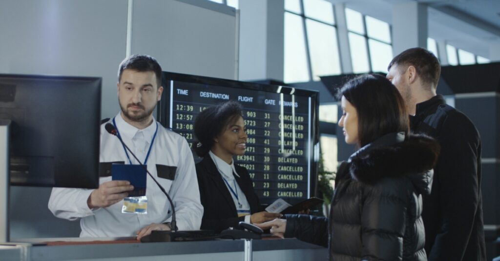 Staff assist two passengers at an airport check-in counter with a departures board showing delays behind them.