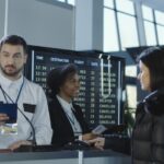 Staff assist two passengers at an airport check-in counter with a departures board showing delays behind them.