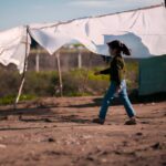 A young child walks past a makeshift shelter made of fabric sheets in a dry, open area.