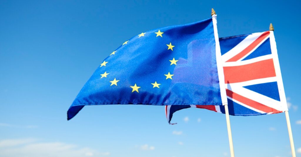 The EU flag and the UK flag wave side by side in the wind against a bright blue sky.