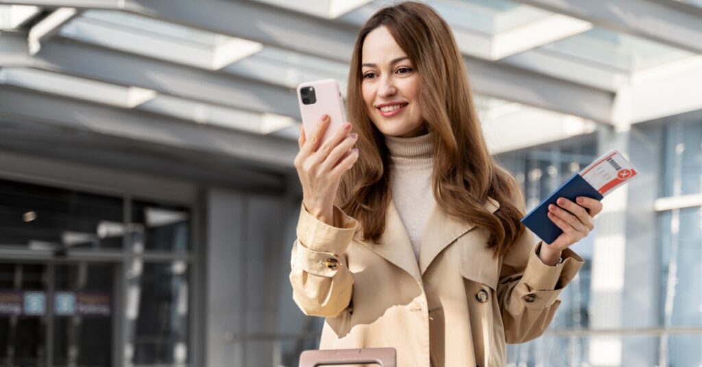 A smiling traveler checks her phone while holding a passport and boarding pass beside her luggage at the airport.