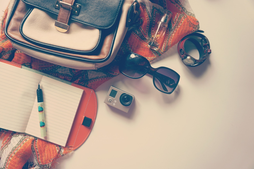 Flat lay of travel essentials including a handbag, camera, sunglasses, notebook, watch, and scarf on a light surface.