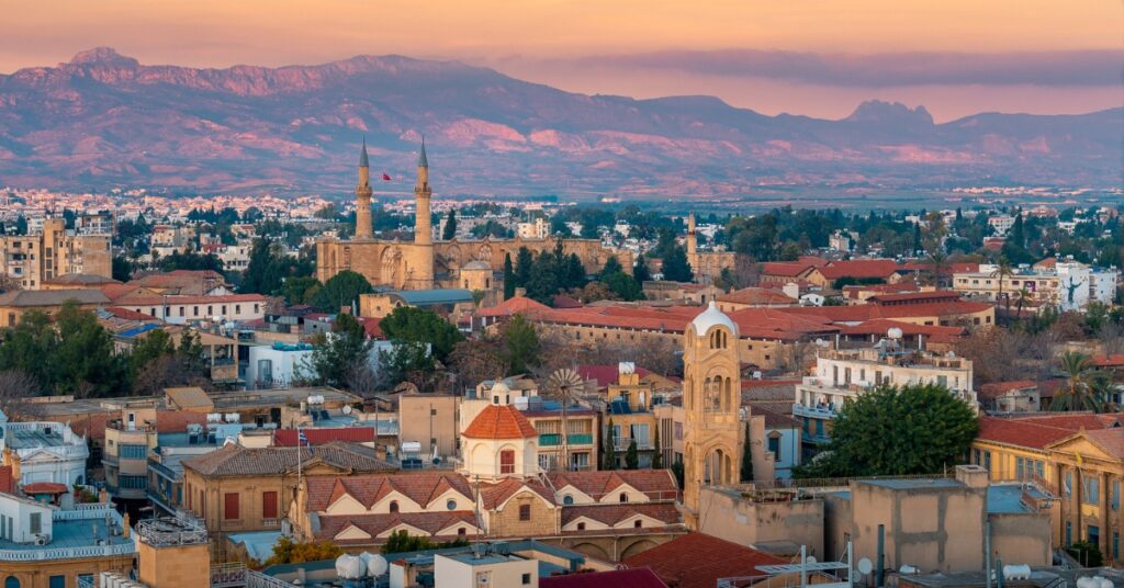 A panoramic view of Nicosia at sunset, featuring historic buildings, church towers, and distant mountains.