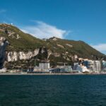 Coastal city skyline with the Rock of Gibraltar rising behind it, viewed from the sea under a clear blue sky.