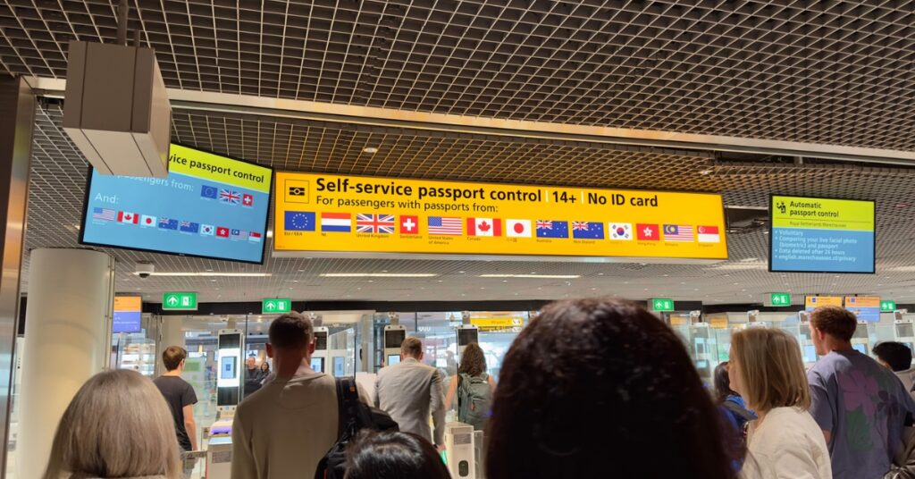 Photograph of travelers queuing at self-service passport control gates beneath overhead signs in an airport terminal.